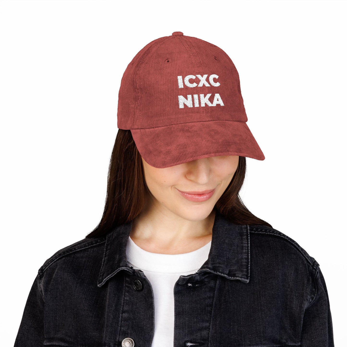 Woman wearing a white ICXC NIKA Christogram cap ("Jesus Christ Conquers")—Orthodox embroidered cotton corduroy—paired with a black denim jacket and white shirt, looking down.