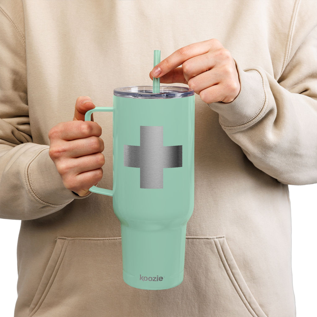 A person in a beige hoodie sips through a straw from the Minimalist Cross Engraved 40oz Koozie Tumbler—a mint green, insulated stainless steel accessory—capturing an everyday moment that contrasts with the solemn Exaltation of the Cross observed by Orthodox Christians.