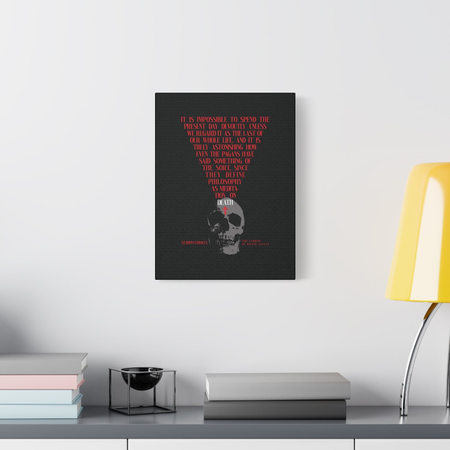 A modern desk with books, a bowl, and a yellow lamp sits beneath "The Present Day As the Last Day of Our Life" Orthodox Canvas Wall Art—featuring a skull and red cross in the memento mori tradition.