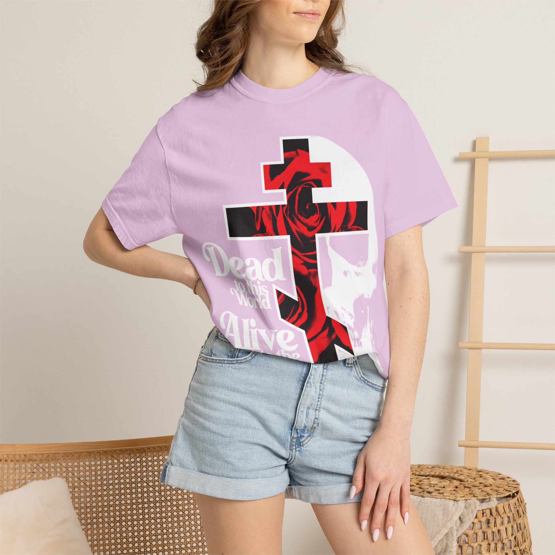 A woman in the "Dead to this World" Christian Goth flower cross & skull graphic T-shirt and denim shorts stands by a wicker chair and wooden rack indoors, showcasing the bestselling heavyweight cotton unisex tee.