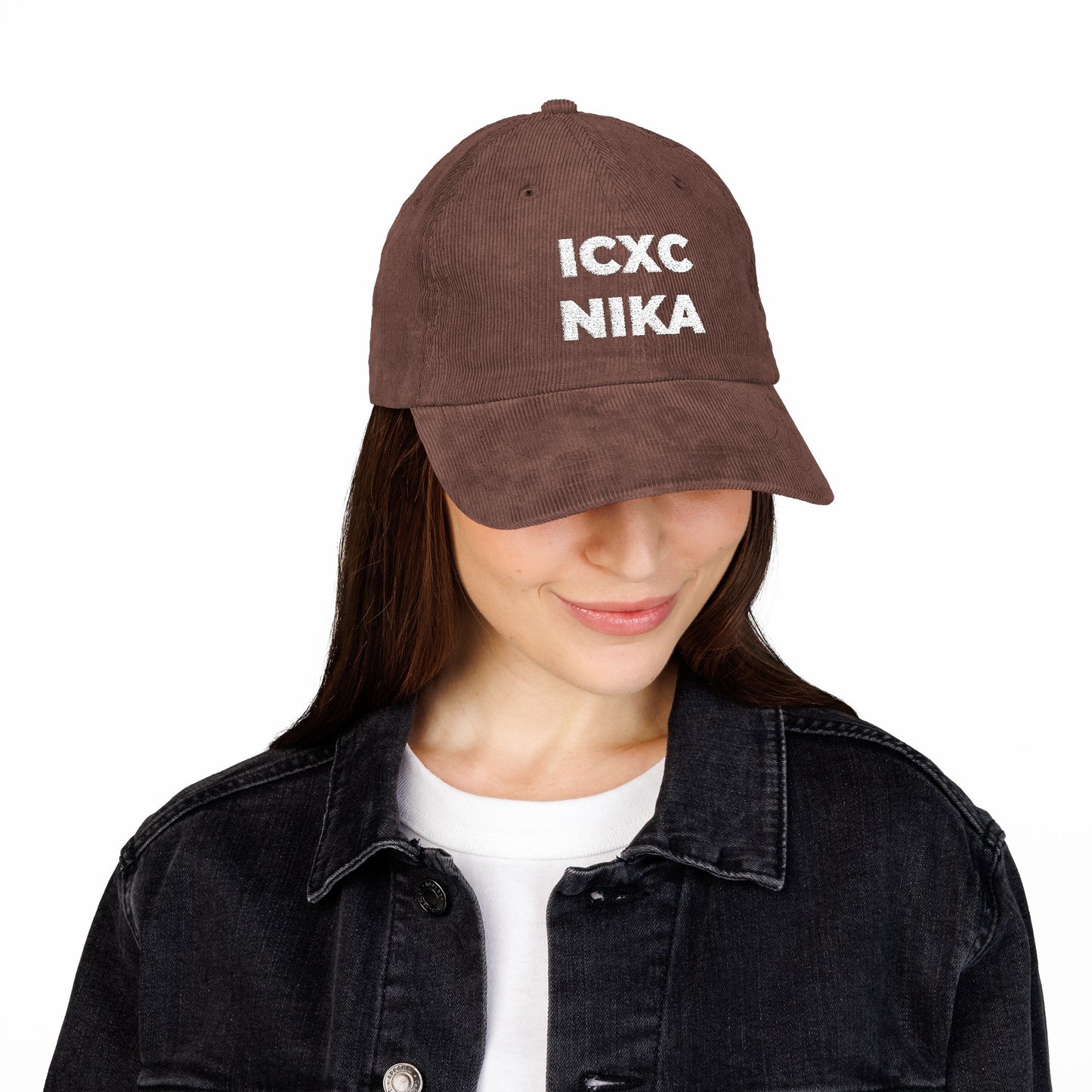Woman in a white ICXC NIKA Text Christogram Orthodox Christian cap—meaning "Jesus Christ Conquers"—embroidered on cotton corduroy, paired with a black denim jacket and white shirt, looking down.