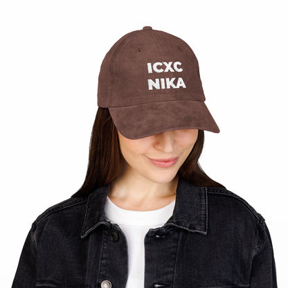 Woman in a white ICXC NIKA Text Christogram Orthodox Christian cap—meaning "Jesus Christ Conquers"—embroidered on cotton corduroy, paired with a black denim jacket and white shirt, looking down.