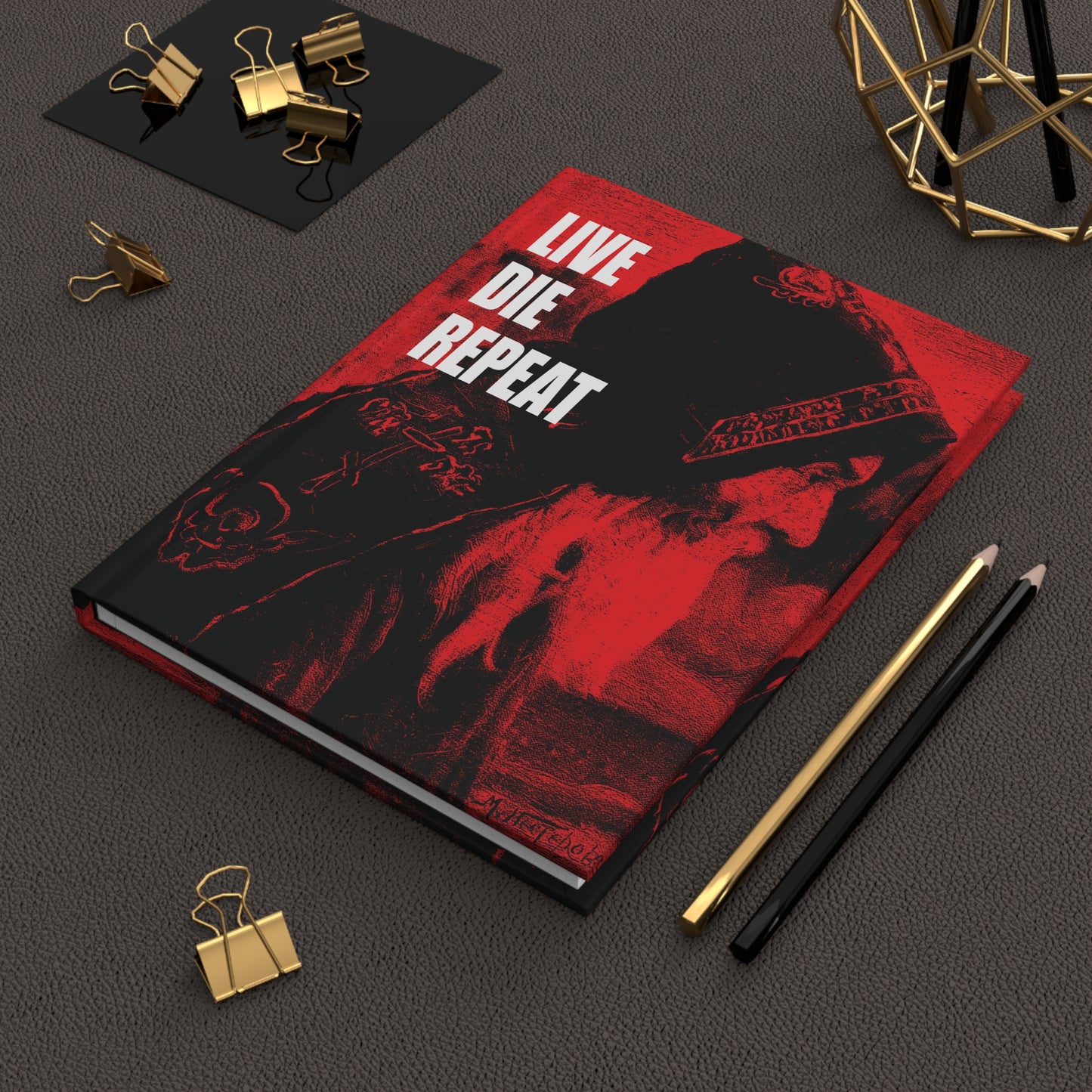 A red and black Live Die Repeat hardcover journal—featuring “Die Every Day, and You Will Live Forever” from Orthodox Christian ascetic teachings—rests on a desk beside pencils and gold binder clips.