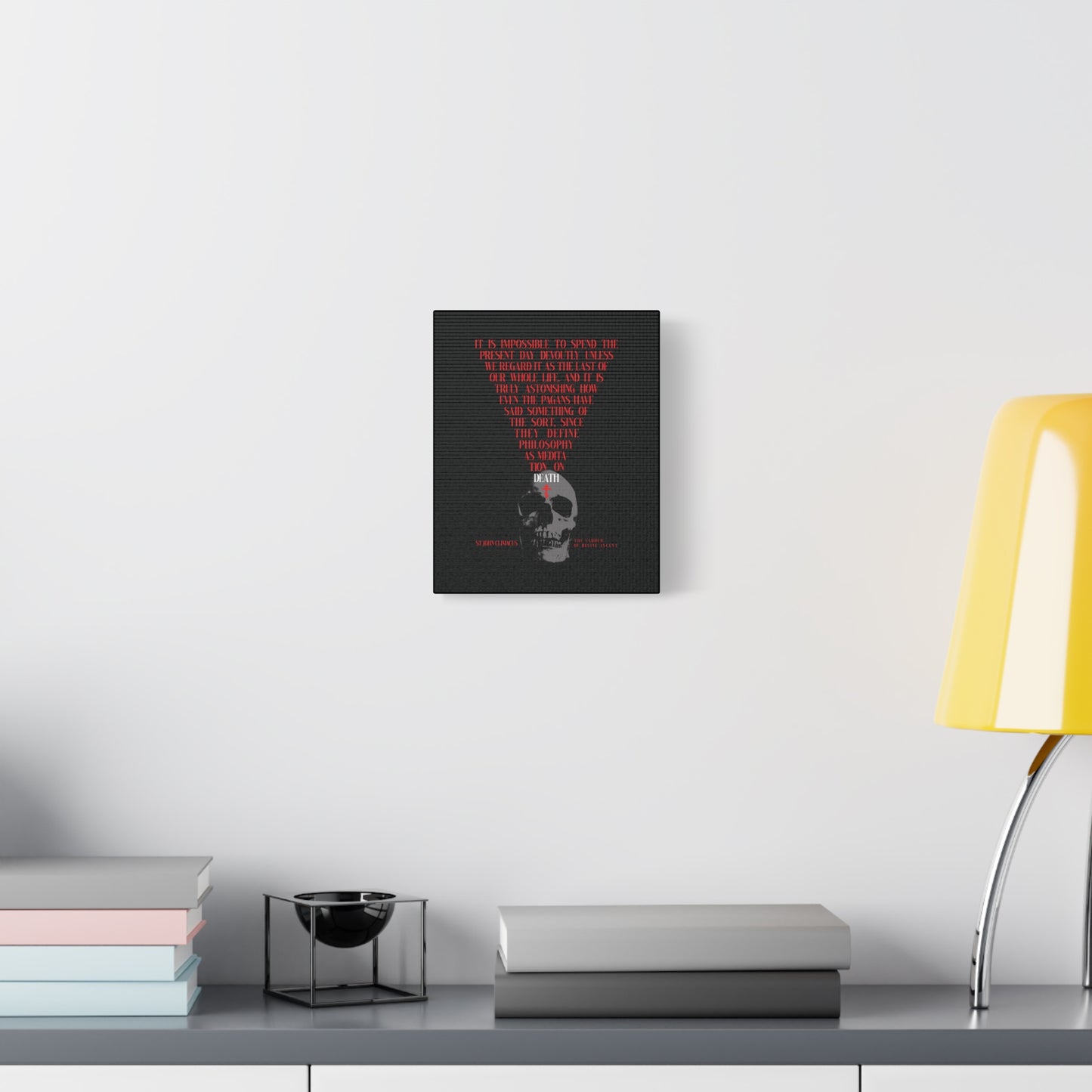 The Present Day As the Last Day of Our Life — an Orthodox canvas wall art featuring a skull with a red cross and memento mori theme — hangs above a desk with books, a bowl, and a yellow lamp on a white wall.