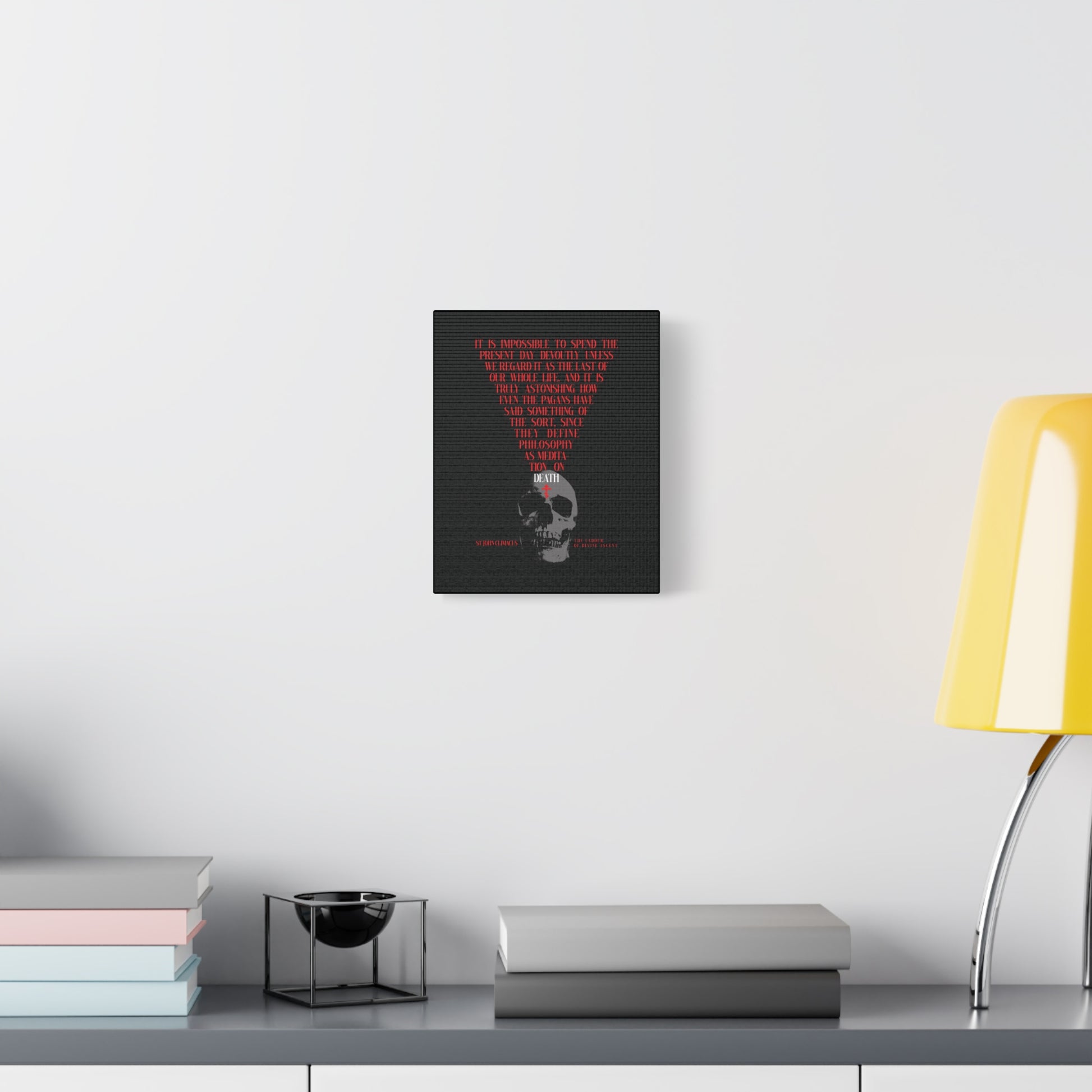 The Present Day As the Last Day of Our Life — an Orthodox canvas wall art featuring a skull with a red cross and memento mori theme — hangs above a desk with books, a bowl, and a yellow lamp on a white wall.