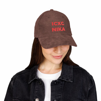 Woman in black jacket wearing the embroidered cotton corduroy cap with red ICXC NIKA text Christogram, meaning "Jesus Christ Conquers," from the Orthodox Christian symbol collection, looking down and smiling.