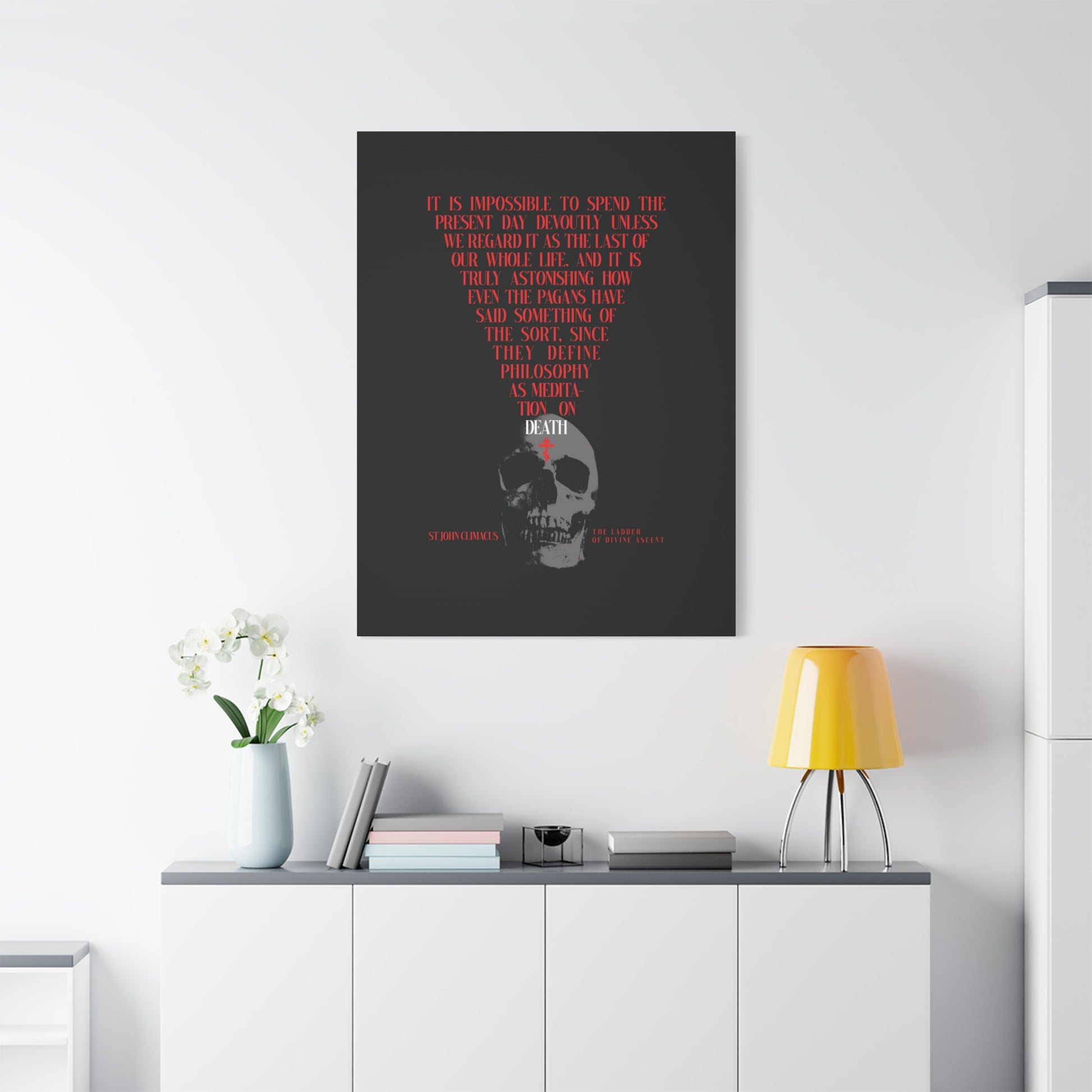 A modern room with a white console, yellow lamp, and flowers features "The Present Day As the Last Day of Our Life" Orthodox canvas wall art—St John Climacus’s memento mori with skull and red cross—inviting reflection on mortality.