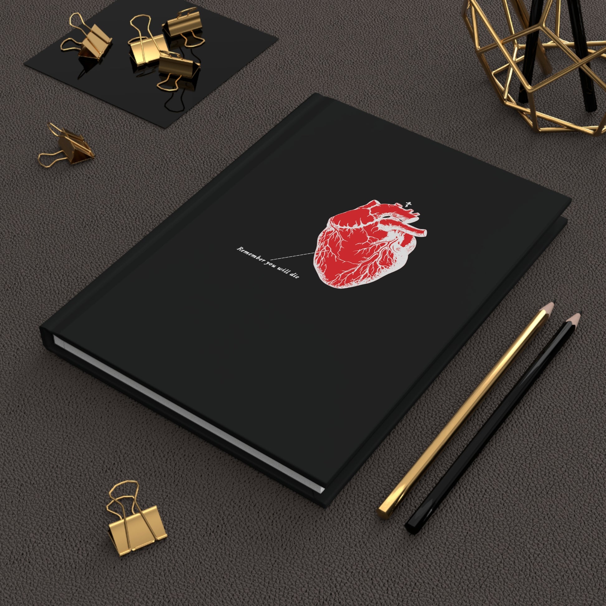 A black Remember You Will Die notebook with a red anatomical heart design and Orthodox Christian quotes on the cover is paired with gold and black pens, gold paper clips, a geometric object, and a small black pad on the dark surface.