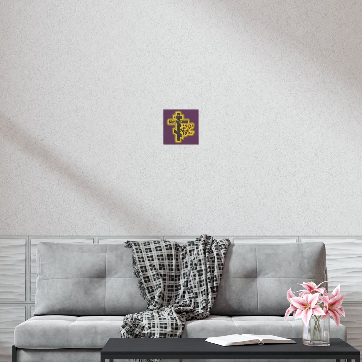 The "Every Knee Shall Bow" Orthodox Christian art poster, with a 70s distressed comic book style, hangs above a gray couch with a plaid throw, creating a peaceful setting completed by a coffee table topped with flowers and an open book.