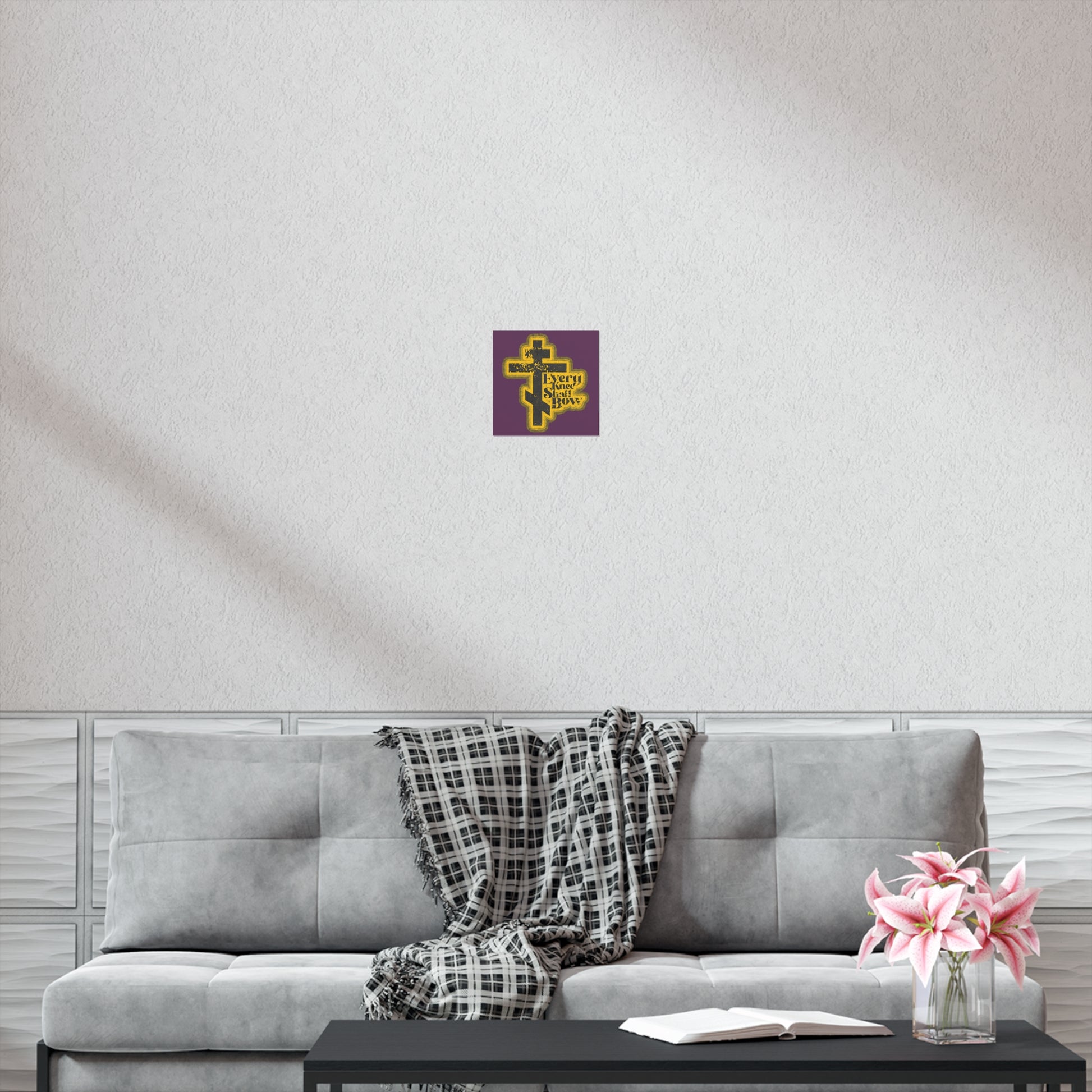 The "Every Knee Shall Bow" Orthodox Christian art poster, with a 70s distressed comic book style, hangs above a gray couch with a plaid throw, creating a peaceful setting completed by a coffee table topped with flowers and an open book.