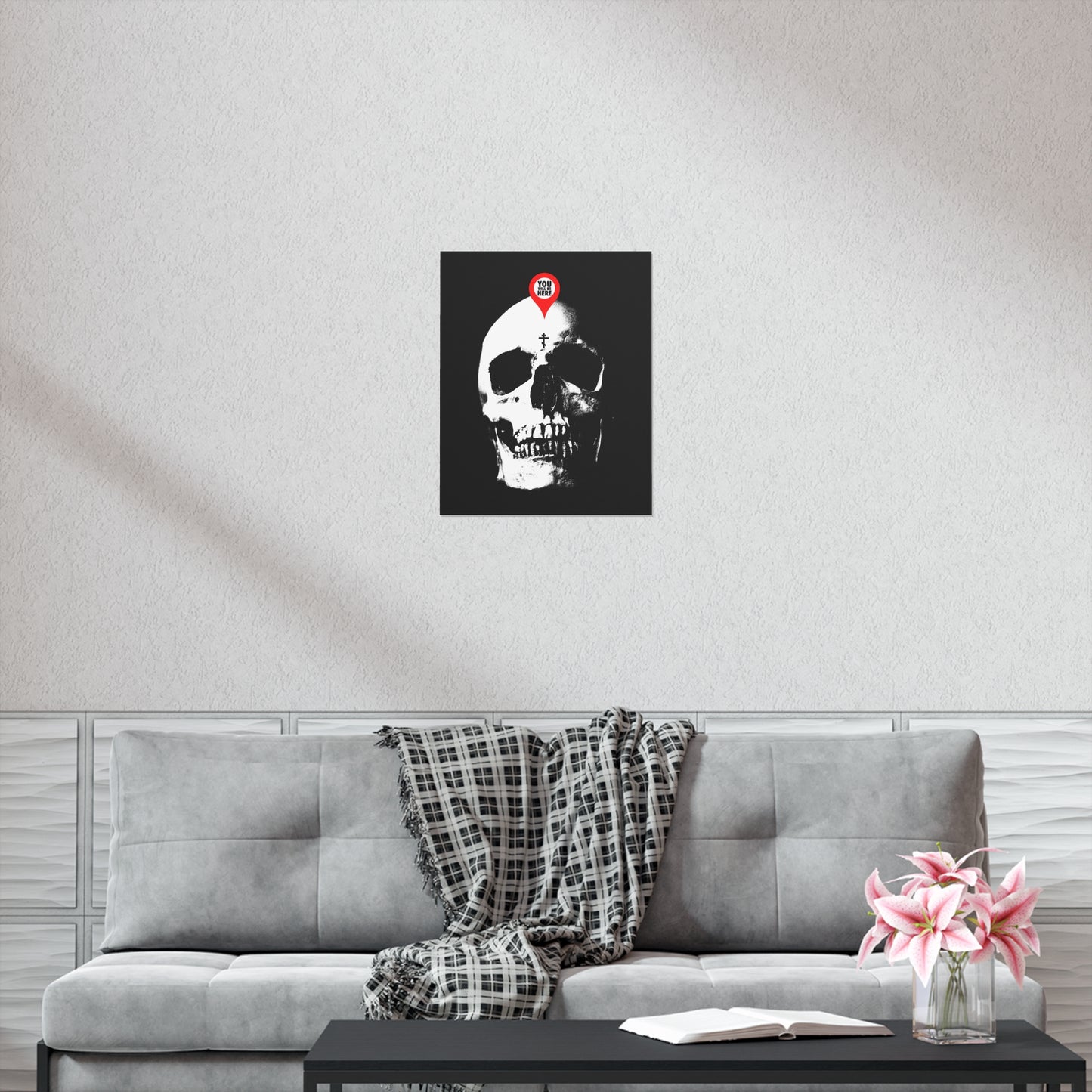 A "You Will Be Here Maps Marker on Skull" Orthodox Christian art poster hangs above a gray sofa; flowers and a book rest on the coffee table, evoking memento mori and reflection on mortality.