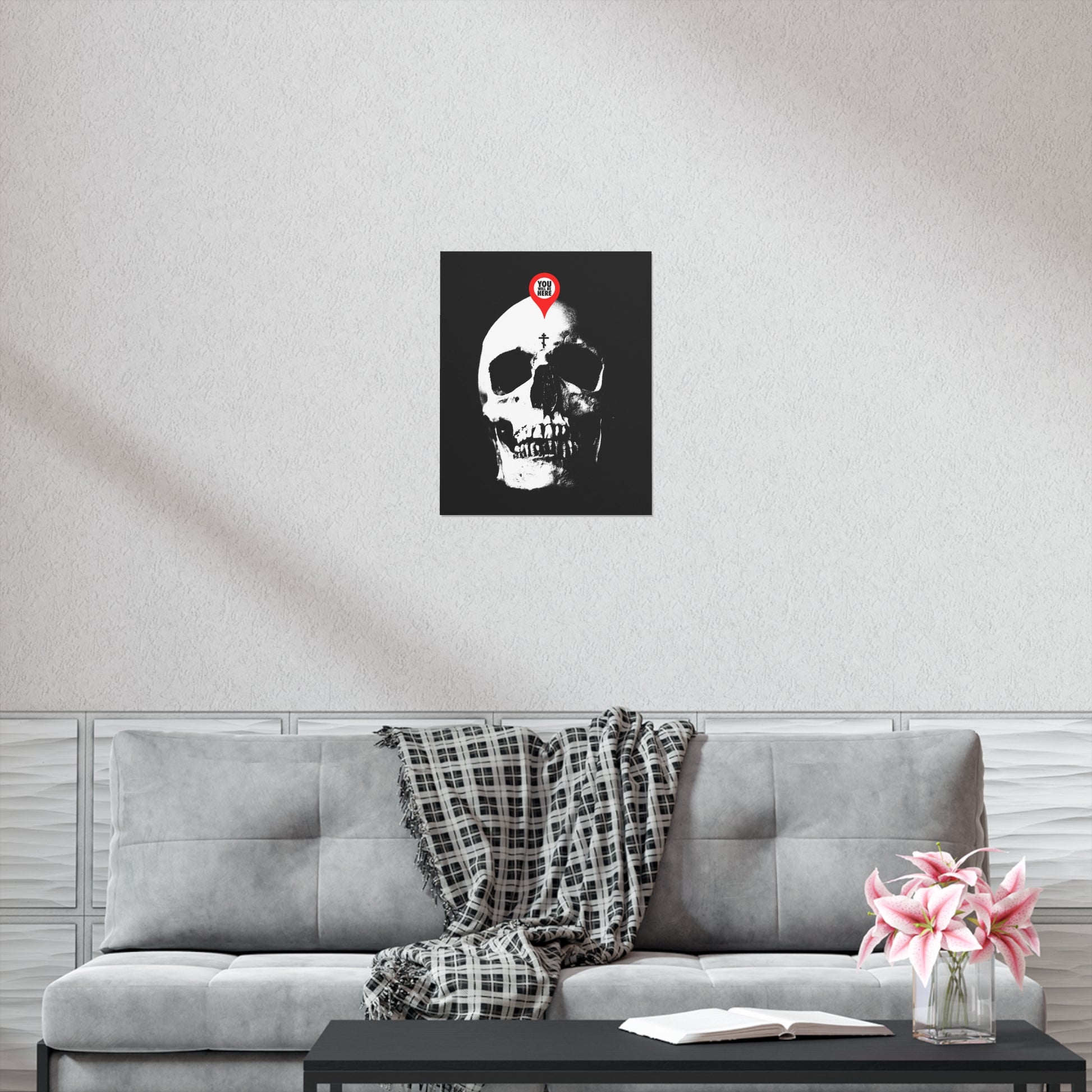 A "You Will Be Here Maps Marker on Skull" Orthodox Christian art poster hangs above a gray sofa; flowers and a book rest on the coffee table, evoking memento mori and reflection on mortality.