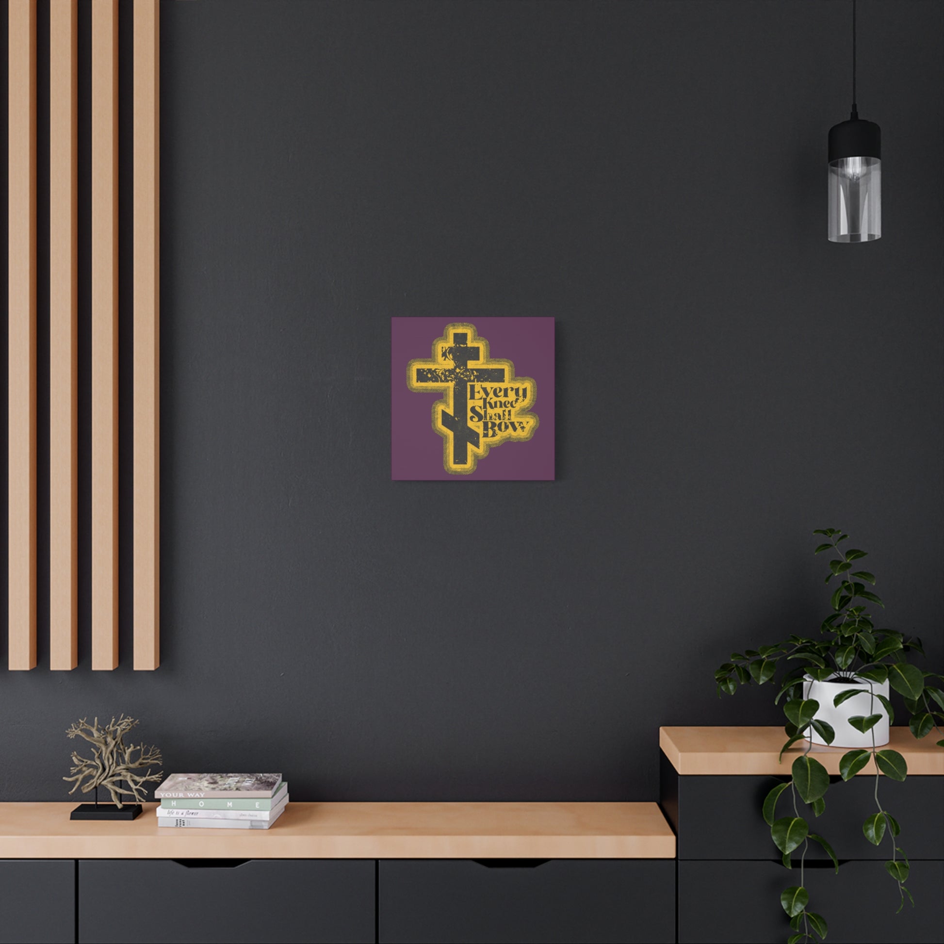 A modern interior with dark walls, plants, books, wooden shelves, and the "Every Knee Shall Bow" Christian wall art featuring a 70s distressed halftone comic book style inspired by Philippians 2:10-11 and Isaiah 45:23.